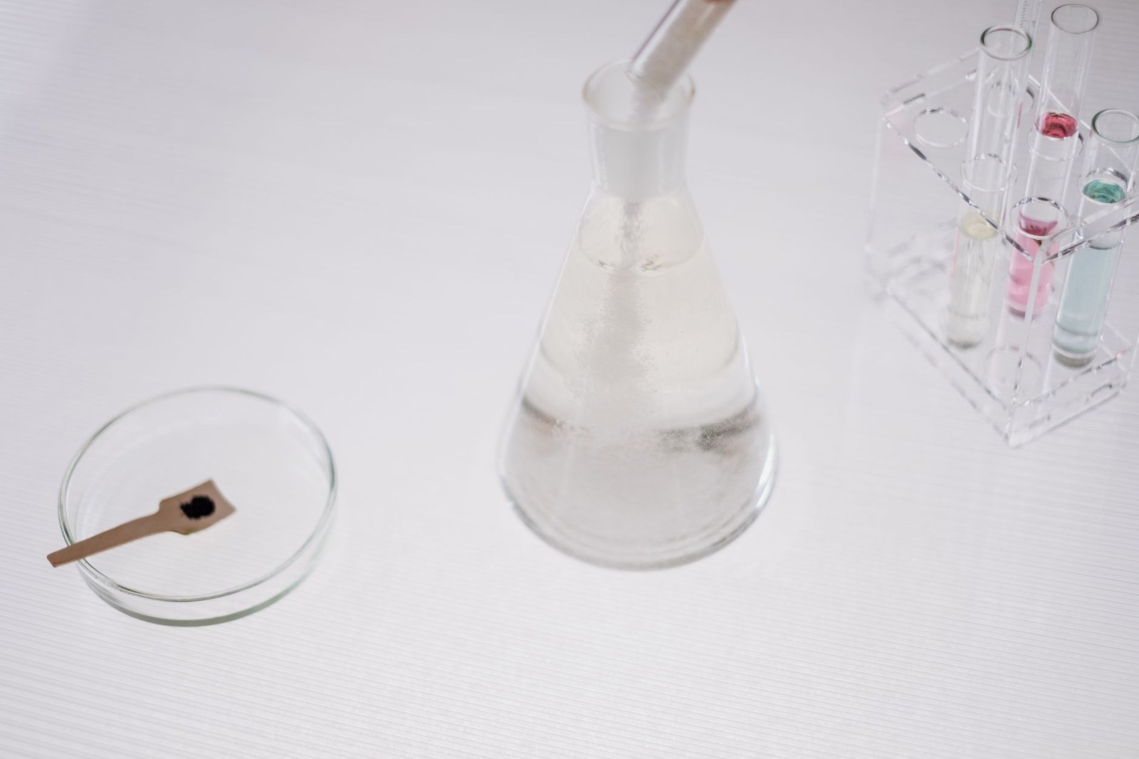Vista aproximada de material de laboratório com um balão, tubos de ensaio com líquidos coloridos e uma placa de Petri com uma espátula contendo pó preto
