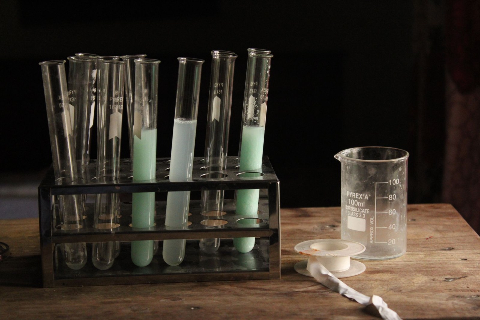 Des éprouvettes contenant des liquides de couleurs claires sur un support et un verre doseur posés sur une table en bois, dans un laboratoire à l’éclairage tamisé.