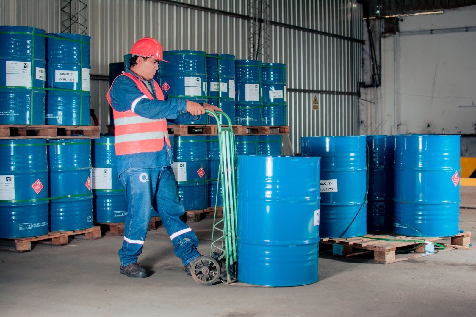 Industrial worker wearing a hard hat and safety vest, moving blue barrels in a warehouse.